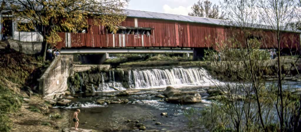 An old red covered bridge over a waterfall, with a small boy in the foreground