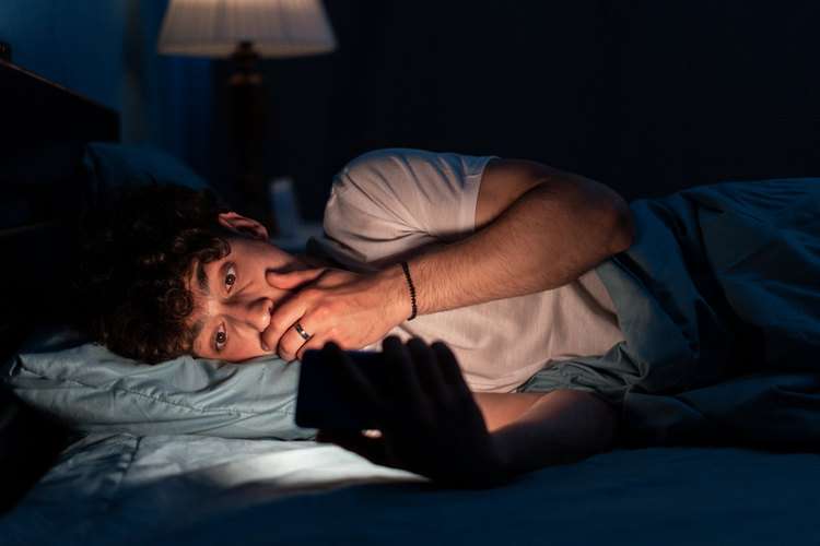Young man lying in bed looking at phone in shock