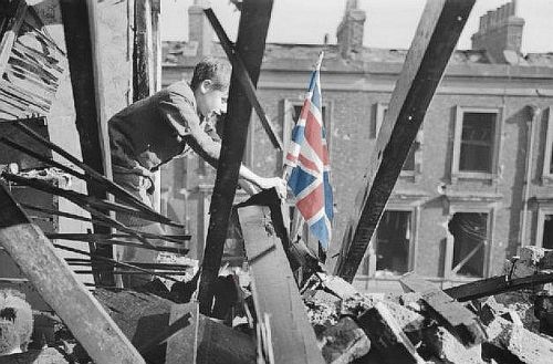 A young smiling boy plants a Union Jack in the midst of rubble